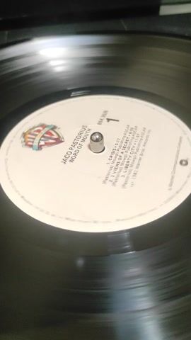 A close-up shot captures a black vinyl record spinning on a turntable. The record's white label is prominently displayed, with a metallic spindle at its center.

The label features the artist's name, "JACO PASTORIUS," and the album title, "WORD OF MOUTH," printed in black capital letters. Below this, the Warner Bros. Records shield logo is visible. To the right of the spindle, the catalog number "BSK 3636" and the side indicator "1" are printed.

A tracklist is also visible on the label:
1.  CRISIS - 5:17
2.  VIEWS OF A SECRET - 6:05
3.  LIBERTY CITY - 11:57

Each track also lists "(Pastorius) Movgli Publishing/ASCAP" as publishing information. At the bottom, "℗ 1981 Warner Bros. Records Inc." is noted. The record is actively playing, indicated by its rotation.