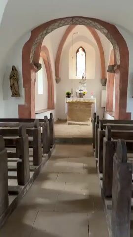 The video shows the interior of a small church or chapel. The perspective is from the aisle, looking towards the altar. There are rows of wooden pews on either side of the aisle. The walls are white, and the arched ceiling has a decorative pattern in red and white. The altar is at the far end, with a stone base and a white cloth covering the top. On the altar, there are religious statues, flowers, and possibly candles. A stained-glass window is visible behind the altar. A statue of a person holding a child is mounted on the wall to the left. The floor is made of stone tiles. The lighting suggests it is daytime, with light coming from the window and possibly other unseen sources. There are no people visible in the video.