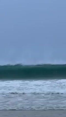The video shows a beach scene with rough waves under an overcast sky. Initially, the video focuses on the large, foamy waves breaking onto the shore. Then, two surfers appear, sitting on their boards and facing away from the camera, riding the waves.