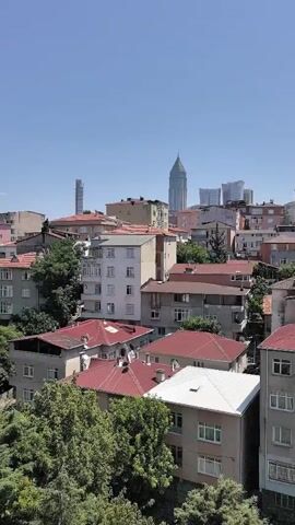A drone shot pans across a dense urban landscape under a clear blue sky. Numerous apartment buildings with red-tiled roofs dominate the foreground, interspersed with lush green trees. In the distance, modern skyscrapers rise, contrasting with the older architecture. The camera moves slowly, revealing more of the city's layout and skyline. No people are visible in the footage. The sun appears to be high in the sky, suggesting it is midday.