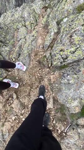 Two individuals stand on a rocky mountain path. The camera's view is initially focused downwards, showing their feet and the immediate terrain - a mix of rocks, small pebbles, and sparse vegetation. One person wears black pants and black shoes, while the other wears black leggings and white and pink sneakers. The camera then tilts upwards to reveal a scenic view of a mountainous landscape with a small waterfall cascading down a rocky cliff. The sky is overcast, with a mix of dark clouds and patches of blue. The mountains are mostly rocky, with some green vegetation scattered across the slopes.