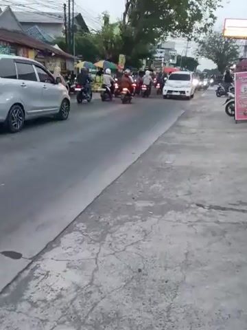 The video shows a street scene with traffic. A white minivan with yellow and red lettering on its side is driving on the road. Several motorcycles are also visible, some with riders and some parked. The road appears to be paved and has some cracks. Buildings and trees line the street. The sky is overcast. The video is filmed from a low angle.
