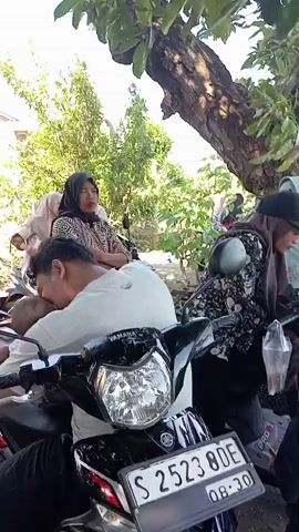 Here's a forensic description of the video:

The video shows a group of people gathered outside in a shaded area, likely a yard or roadside location. Several motorcycles are parked nearby, some with people sitting on them. The adults mostly wear traditional clothing, including hijabs. One man in a light-colored shirt is sitting on a motorcycle, with his head resting on the vehicle, appearing tired. A child in a black t-shirt is seen near the front motorcycle, looking at it. In the background, another man in an orange shirt is sitting on a wall with his feet on a motorcycle. The ground is uneven and appears to be a mix of dirt and some greenery. Several trees provide shade.