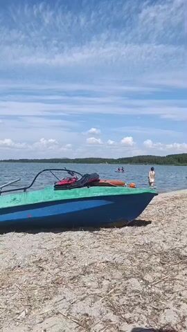 A blue and green boat is partially visible on a sandy beach. The boat has a red object and some bags in it. The water is calm and there are a few people in the water. The sky is blue with some white clouds. In the distance, there are trees and hills. The video pans to the right to show more of the lake and the wooded hills.