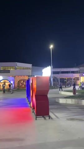 A vertical shot shows a nighttime outdoor scene with a large, illuminated sign that reads "BUILD EX". The letters are lit with purple, blue, yellow, and red lights. The sign is in the foreground, and behind it, there are buildings with windows and lights, streetlights, and a few people walking around. The ground is paved. The sky is dark.