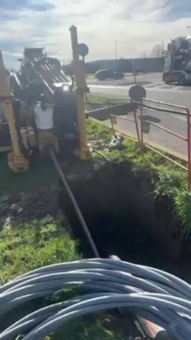 The video shows a yellow drilling machine next to a rectangular hole in the ground. A long pipe is entering the hole, presumably being drilled underground. Coiled gray cables are visible in the foreground. A street and traffic can be seen in the background.