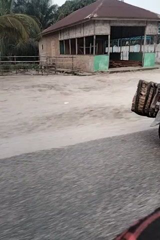 The video shows a person riding a motorcycle with a large woven bag on the back. The motorcycle is moving down a paved road. There are buildings and trees in the background. The camera is moving and appears to be handheld.