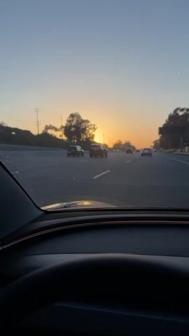 The video shows a view from inside a car, looking out through the windshield. The car is on a highway, and the sun is setting in the distance, casting a warm glow across the sky and the road. Several other cars are visible on the highway, driving in the same direction. The road appears to be asphalt, with clear lane markings. Trees and vegetation line the sides of the highway. The sky is a gradient of blue and orange, indicating the time of day is dusk. The interior of the car, including the dashboard and steering wheel, is partially visible in the foreground. The overall scene is one of driving on a highway during sunset.