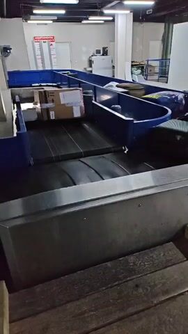 The video shows luggage moving on a conveyor belt. The conveyor belt is set in a blue frame and is transporting a cardboard box, bags, and a suitcase. The conveyor belt is located inside a large, well-lit room that may be an airport or warehouse. The room contains a white door, a desk, and a sign. The camera is slightly off-center and at a low angle.
