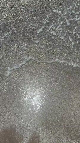 The video shows a close-up view of the ocean waves rolling onto a sandy beach. The water is clear and reflects the sunlight, creating a shimmering effect on the wet sand. The waves are gentle, with white foam forming as they break. The sand appears to be made of small pebbles and grains. The camera is positioned at an overhead angle, looking down at the water and sand. The video is very short, lasting only a few seconds, and captures the natural beauty of the beach.