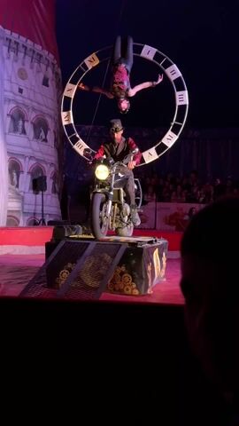 A circus act features a person dressed in a steampunk-style outfit sitting on a motorcycle, poised on a small platform with ramps. Above them, another person hangs upside down within a large circular structure resembling a clock face. The stage is set with a red floor and decorated with gear motifs. In the background, there's a backdrop of what appears to be a stylized building façade. An audience is visible in the background, and a person's head is slightly visible in the foreground, suggesting the viewpoint is from within the audience. The lighting shifts through different colors, adding to the theatrical atmosphere.