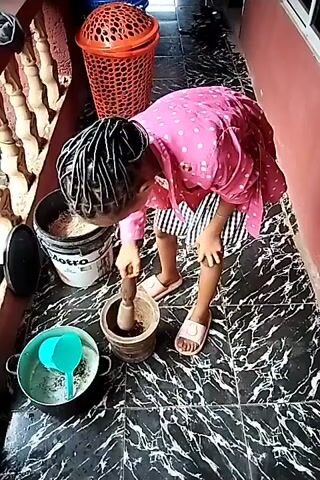 The video shows a child with braided hair, wearing a pink polka dot shirt and black and white striped shorts, grinding something in a mortar with a pestle. The child is standing on a black and white marbled tile floor on what appears to be a porch. Near the child are a bucket, a pot with a blue measuring cup inside, and two lidded wicker baskets of different colors. The porch has a red wall on one side and a white railing on the other. The child is focused on the grinding task, pressing the pestle into the mortar with both hands.