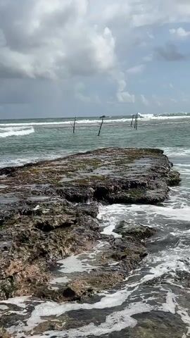 A scenic view of waves crashing onto rocks in the ocean. In the background, there are several stilts or poles sticking out of the water, and the sky is cloudy with patches of blue. The waves are breaking against the rocky shore, creating foam and spray. The overall scene is one of natural beauty and the power of the ocean.