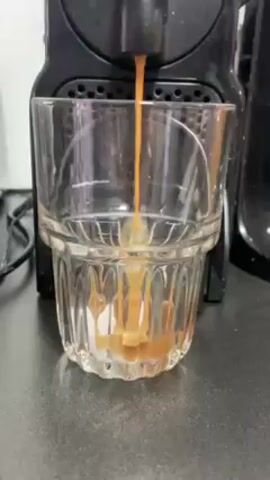 A small, black, Nespresso coffee machine brews coffee into a clear, faceted glass. The coffee is a light brown color and fills the glass to approximately one-third full. The counter on which the machine and glass are placed is black. The environment appears to be indoors.