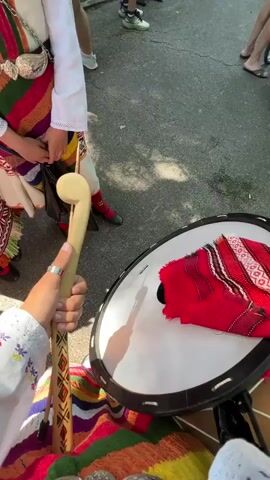 A person in a colorful traditional costume is playing a large drum with a wooden stick in a park setting. Another person is also visible in the left foreground, possibly playing another instrument or observing. In the background, there is a paved pathway with numerous people walking around and observing, some standing in groups. The environment is a park with many green trees, suggesting an outdoor event or festival. There are also some stalls or tents visible in the distance, further indicating a public gathering. The weather appears to be sunny and pleasant.