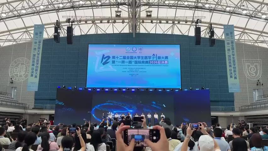 A wide shot shows a large indoor arena with a crowd of people seated in the foreground, facing a stage. Several people are holding up smartphones to record. On the stage, a large LED screen displays text and graphics. Above the LED screen, a banner reads in Chinese "The 12th National Undergraduate Medical Innovation Tournament & The Belt and Road International Competition 2026 Finals". Below this, another banner is partially visible. A group of individuals, likely contestants or organizers, stand on the stage, facing the audience. The arena has a high, arched ceiling with exposed structural elements and hanging speakers. Another large screen is positioned above the main LED screen. The overall atmosphere appears to be that of a formal event or competition.
