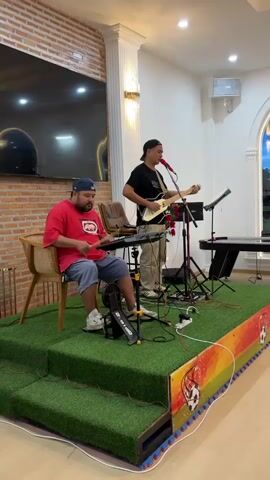 A man in a red shirt and blue shorts sits on a chair playing drums while another man in a black shirt and beige pants plays a guitar and sings. They are on a raised stage covered in artificial grass, decorated with soccer balls. In the background, there is a brick wall and a large television.
