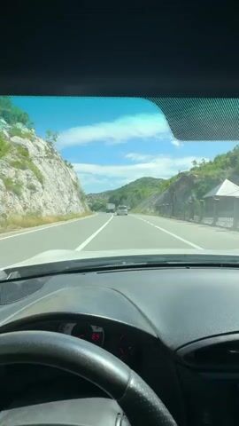 A first-person perspective video from inside a vehicle traveling on a highway. The vehicle is moving forward on a paved road with a white line marking the lane. The dashboard and steering wheel are visible in the foreground. The road is flanked by rocky hillsides and green vegetation. The sky is blue with scattered white clouds. Other vehicles, including a truck and a smaller car, are visible ahead on the road. Traffic signs are present on the right side of the road. The time code indicates the video is approximately 5 seconds long.