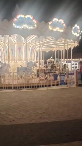 A carousel at night with bright lights. The carousel has white horses. There are people around the carousel, some entering and exiting. The ground is paved with interlocking stones.