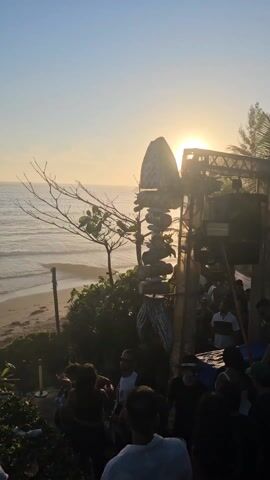 The video shows a vibrant beachside event during sunset. The sky is clear with the sun low on the horizon, creating a golden glow. The sea is visible in the background with small waves. In the foreground, there's a crowd of people gathered near a structure possibly a stage or bar area. A unique sculpture made of stacked stones stands prominently, backlit by the sun. The event seems lively, with music likely playing. There are plants and greenery, adding to the natural setting.