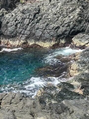 The video shows a small cove surrounded by dark, jagged rocks. The water in the cove is a vibrant turquoise color, with white foam swirling on the surface as small waves enter from the side. The rocks are rough and uneven, with various shades of gray and black. The perspective is a high-angle shot looking down into the cove. The camera remains static throughout the video, capturing the natural movement of the water and the rugged texture of the surrounding rocks.