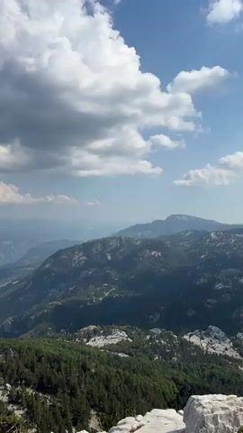The video displays a panoramic view of a mountainous landscape under a partly cloudy sky. The foreground shows the rocky terrain and sparse pine trees. In the middle ground, rolling hills covered with dense green forests are visible, with some patches of lighter rock formations. The background reveals a series of distant mountain ranges fading into a hazy blue. The clouds are white and puffy, with some darker gray clouds present, suggesting dynamic weather. The sun appears to be shining, casting shadows on the mountains. There are no people or man-made objects visible in the video. The overall impression is one of a vast, wild, and natural environment.