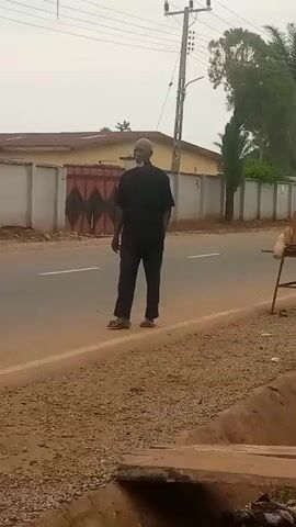 The video shows an elderly man with a beard, wearing a black shirt and pants, walking on the side of a road. He is on a gravelly shoulder next to a paved road. Behind him, across the road, is a building with a tan-colored facade and a gray wall. There is a large gate with a diamond pattern on the building's wall. A utility pole with wires stands near the building. A woman wearing a patterned outfit is also walking on the road, behind the man. The scene appears to be outdoors during the day, with overcast skies. The setting suggests a rural or semi-urban area in Nigeria, as indicated by the architecture and the utility pole. The man's gait suggests a relaxed pace. There is no visible text in the video.