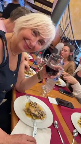 This is a close-up, slightly over-the-shoulder shot of a smiling woman with short, blonde hair and bangs. She is wearing a denim top with buttons down the front and a gold necklace. She is holding a glass of red wine in her right hand, bringing it closer to the camera. Her left hand is holding a knife and fork over a plate of food, which appears to be seafood in a sauce. The plate is white with a gold rim and is set on a table covered with a red tablecloth.

In the background, other people are seated at tables, also appearing to be dining. One woman with brown hair is visible, wearing a light brown top. Another person is partially visible in the upper left corner, wearing a pink shirt. There are bottles of water and wine on the table. The setting appears to be a restaurant, suggested by the table setting, the presence of multiple diners, and a glimpse of a staircase in the upper right corner. The lighting suggests it is daytime, likely indoors. The woman's smile and the raised glass of wine imply a celebratory or convivial mood. The food on the plate suggests a meal is in progress.