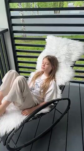 Here's a description of the video:

The video shows a young girl sitting in a black rocking chair on a porch. The chair is covered with a fluffy white cushion. She is wearing a white t-shirt and light beige pants. She appears to be relaxed, occasionally looking around. The porch has a black deck and black slatted railing. In the background, there is a view of greenery, trees, and a white fence. The weather seems overcast.