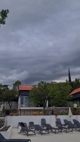 A low-angle shot shows a row of empty lounge chairs by a swimming pool. In the background, modern buildings with red tile roofs and white walls are nestled among trees. The sky is filled with dark, ominous storm clouds. A person in a green shirt is standing on a stone wall, and another person in a white shirt is visible on a balcony.