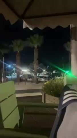A night-time video shot from a seated position under a light-colored parasol reveals a view of a partially-illuminated area with palm trees and parked cars. The person filming is wearing blue and white striped slides. A man dressed in a black jacket, light-colored shorts, and dark sandals walks from left to right across the view, pausing briefly before continuing out of the frame. There is a green laser dot visible in the scene.