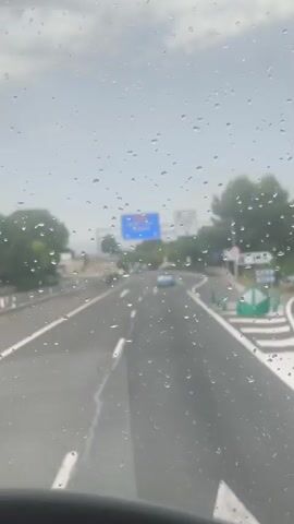 The video captures a view from inside a vehicle traveling on a highway in Llíria, Spain. The scene is dominated by the road ahead, marked with white lane lines and arrows, and viewed through a windshield covered in raindrops. The weather appears to be overcast and rainy.

On the highway, several vehicles are visible, including a light-colored car in the distance. Traffic signs are present, with a prominent blue sign indicating "CV-35 Llíria (oest) Ademuz" with downward arrows, suggesting directions to these locations. Another white sign on the right reads "LLIRIA (nord)" with a downward arrow. A speed limit sign indicating "70" is also visible.

The surrounding landscape consists of trees and foliage lining the highway, with some signs of distant hills or mountains. The overall mood is subdued due to the weather and the perspective from within the vehicle. The time of day appears to be daytime, but the overcast sky obscures direct sunlight. The presence of raindrops on the windshield and the wipers occasionally sweeping across the view are notable details that emphasize the rainy conditions.
