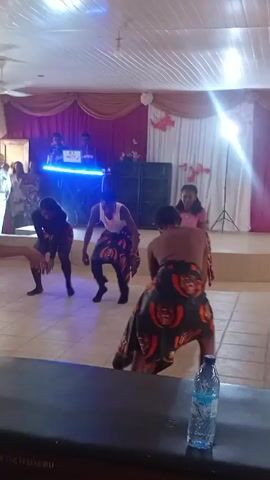 Inside a decorated room, a group of people are dancing to music. Three topless men and two women in dresses, are wearing dark skirts patterned with red and brown faces. The dance involves bending at the waist, shaking their bodies, and moving rhythmically. In the background, two DJs are at a table lit by blue lights. A table with a water bottle is in the foreground. The room has a tiled floor, decorative curtains, and speakers.  The room is lit by fluorescent ceiling lights and a bright light behind the DJs.