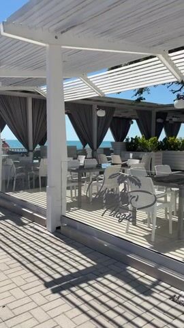 The video shows a view from a paved walkway towards a beachside cafe. The cafe has white canopies with parallel slats and is furnished with white tables and chairs. Gray curtains hang from the sides of the canopies. Through an opening in the cafe, a person in an orange shirt and blue shorts is visible standing on a wooden deck near the sea. The sea is calm and blue, and the sky is clear. The sun casts shadows from the cafe's structure onto the paved walkway.