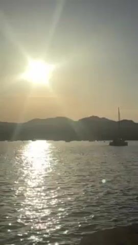 Here is a concise description of the video:

The video shows a scenic view of a body of water with boats silhouetted against the setting sun. The water reflects the sunlight, creating a shimmering effect. In the background, there are mountains. The light in the sky is bright.