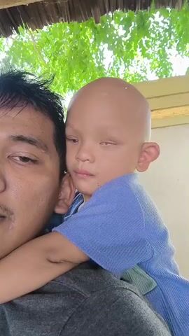A close-up, handheld shot shows a man with dark hair and a mustache, partially visible from the left side of the frame. On his shoulder, a young child with a bald head and wearing a blue shirt is leaning against him. The child's face is turned towards the camera. The background features green foliage and a light-colored building structure. The scene appears to be outdoors, possibly in a tropical setting, with natural light.
