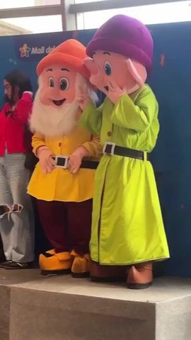 Two individuals dressed as Disney's Seven Dwarfs stand on a platform. On the left is a character with a long white beard, wearing an orange hat, yellow shirt, and maroon pants. The character to the right is wearing a purple hat and a lime green robe. They both have black belts with silver buckles. The character in green holds up two fingers. To the left of the characters is a woman with long dark hair wearing a red shirt and blue jeans. Behind the characters is a blue backdrop with confetti designs and the words "Mall del Sur". The floor appears to be made of gray tile.