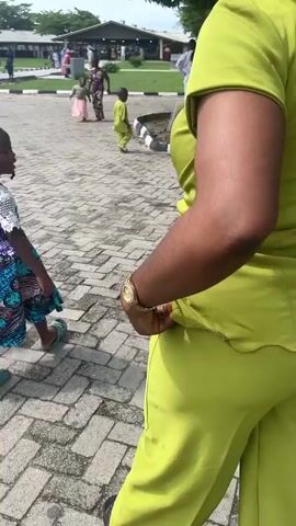 A video shows the back of a woman wearing a lime green outfit consisting of a short-sleeved top with a zipper down the back and flared pants. She has dark, voluminous hair styled in large puffs. In the background, a group of people, including children in colorful dresses, are walking on a paved area. The environment appears to be an outdoor setting with trees and buildings visible. The camera angle is from behind the woman, focusing on her and the activity around her.