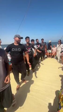 A group of young men are dancing in a line on the deck of a boat. The man in the foreground on the left is wearing a grey t-shirt and black pants with a yellow design. He has his arms raised. The man next to him is wearing a black t-shirt with "D&G" written on it in white glitter and black pants. He is also dancing with his arms raised. The men behind them are also dancing, some in black t-shirts with designs and black pants. On the right side of the boat, a group of people are sitting and watching the dancers. The sky is blue and clear, and the water is visible in the background. The deck of the boat is a light color, possibly wood.