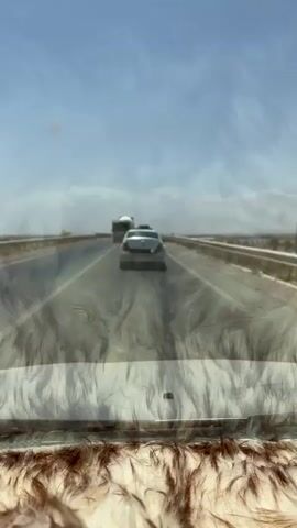 A car passenger filmed a white Renault saloon with its rear trunk door open being driven along a highway under a clear blue sky, with a concrete truck ahead of it. The Renault's Moroccan number plate reads -18366-I-72-. The dashboard of the filming vehicle is covered in fluffy brown material.