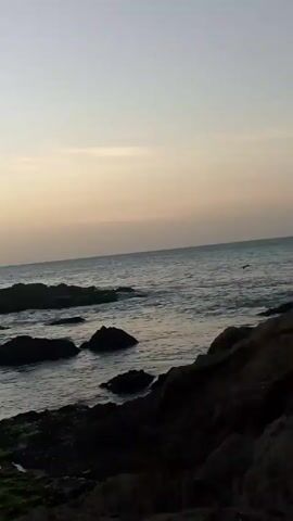The video shows a rocky shoreline meeting the ocean under a pale sky. The scene is dominated by a pile of large, dark-colored rocks in the foreground. Beyond the rocks, the ocean stretches to the horizon. The sky is light, with a gradient of color suggesting either sunrise or sunset. A small boat can be seen in the distance. There are no people visible.