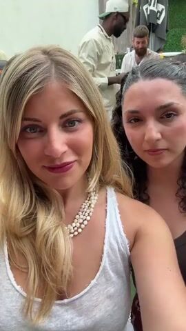 Two young women are taking a selfie outdoors at a casual gathering. The woman on the left has long blonde hair, a white tank top, and a multi-strand pearl necklace. The woman on the right has dark curly hair and a brown top. Both women are wearing makeup. In the background, several people are standing around, chatting. One person is wearing a tan jacket and black sunglasses. There are plants visible and what appears to be a section of a blue cushion. The selfie is taken at an angle and then rotates.