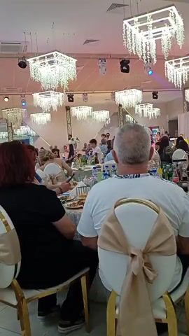 The video shows a crowded indoor event, likely a wedding reception or banquet, viewed from behind several seated guests. The room is decorated with multiple square, tiered chandeliers with cascading crystal-like elements. The walls appear to be mirrors, reflecting the scene. Several tables are set with white tablecloths, plates, and bottles of beverages. In the background, people are seated at tables, some are standing and interacting, and a person appears to be performing on a raised platform or stage. The lighting is a mix of ambient overhead lights and colored stage lights, creating a festive atmosphere. The guests are dressed in casual to semi-formal attire. The focus is primarily on the backs of the heads and shoulders of the guests in the foreground, with the event unfolding in the mid-ground and background.