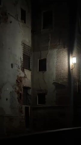 The video shows a dimly lit, weathered brick building at night. A single lantern affixed to the corner of the building casts a warm light on its facade. The building has multiple windows, some of which are barred. The plaster on the walls is peeling, revealing the brickwork beneath, and the scene appears to be an alleyway or narrow street. The lighting suggests a low-light environment, with deep shadows obscuring much of the area. There are no people or animals visible in the video.