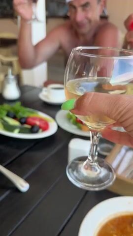 This is a close-up video of a group of people toasting with wine. In the foreground, a hand with green nail polish holds a glass of white wine. In the middle ground, two other hands hold glasses of red wine, and another hand holds a glass of dark red wine. In the background, a man wearing a white baseball cap and no shirt smiles and raises a glass of dark red wine. The setting appears to be an outdoor restaurant with a dark wooden table.