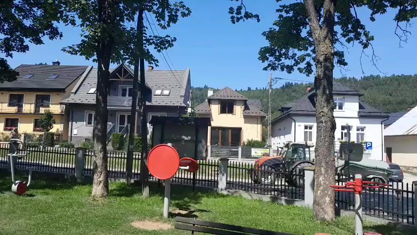 A daytime, outdoor, eye-level shot shows a residential street with several houses and trees. In the foreground, there is a park with exercise equipment and a bench. A black metal fence separates the park from the street. Behind the fence, there are two tractors, one green and one red. The houses are varied in style and color, with some having tiled roofs and others asphalt shingles. The sky is clear and blue, and the sun is shining, casting shadows on the ground. Trees with green leaves line the street, and a forested hill is visible in the background. A white car and a dark-colored car are parked on the side of the street.