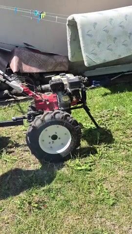 A red and white rototiller is parked on a grassy area next to a building. A young boy wearing a black and red shirt, blue shorts, and yellow Crocs is sitting on a metal seat attached to the rototiller. An adult male, wearing a blue t-shirt and tan pants, walks towards the rototiller from the left side of the frame. He appears to be adjusting his hair. The boy looks towards the camera and then to the right. There are clothes hanging on a line behind the rototiller, and various objects are stacked near the building.