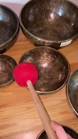 A red mallet with a wooden handle is shown striking a Tibetan singing bowl. Several other Tibetan singing bowls of varying sizes are arranged around it on a wooden surface. The video captures the moment of impact and the reverberation of the bowl.
