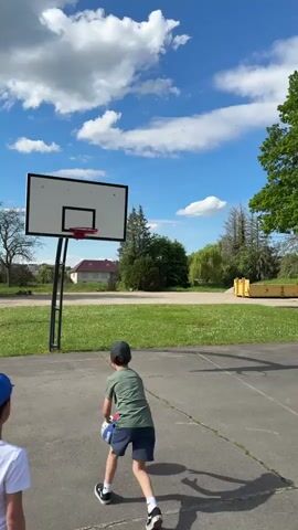 The video shows a young boy on an outdoor basketball court attempting to shoot a basketball into the hoop. He is wearing a green shirt, blue shorts, and a dark hat. The court is surrounded by grass, trees, and a building in the background. The sky is blue with scattered clouds. The boy throws the ball, it hits the rim, and bounces back to him. Another child in a blue cap is partially visible in the lower left corner of the frame. A yellow dumpster is visible in the background.