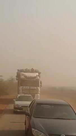 The video shows a rural road scene with very poor visibility due to heavy dust. Three vehicles are visible: a large white truck heavily loaded with what appears to be bags is in the center, followed by a silver car. A black car is parked or moving slowly on the right side of the road. To the left, there's another white truck, possibly a tanker, parked on the shoulder. The road is paved but has dirt on it, and the shoulder appears unpaved. Some scrub vegetation is visible on the sides of the road. The overall atmosphere suggests a dusty or sandy environment, likely a dry, rural area.